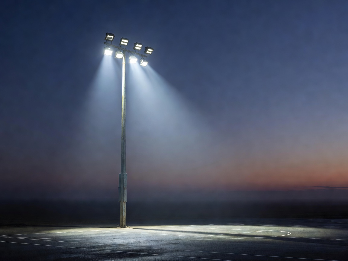 LED floodlight luminaires mounted on a tall sports mast illuminating a court at dusk